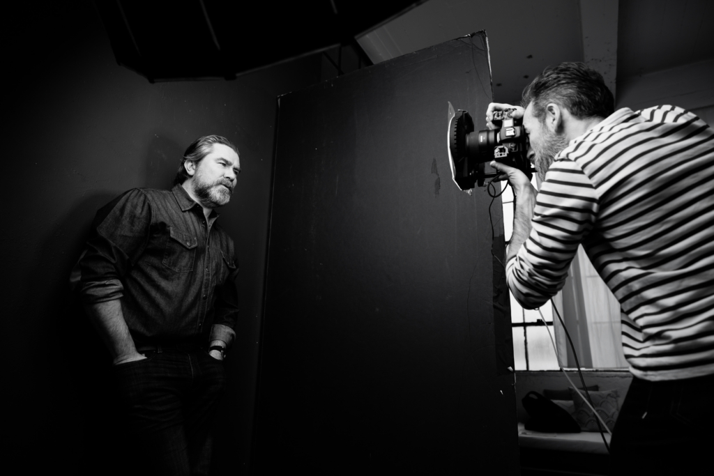 A black-and-white behind-the-scenes photograph of Ivan Weiss working in his studio, leaning forward as he photographs a man standing against a dark wall. A large softbox light hangs above them, shaping the light across her face while he looks through the camera with focused concentration. The scene captures the quiet intensity of a portrait session in progress.

Shot by Rich Soublet.