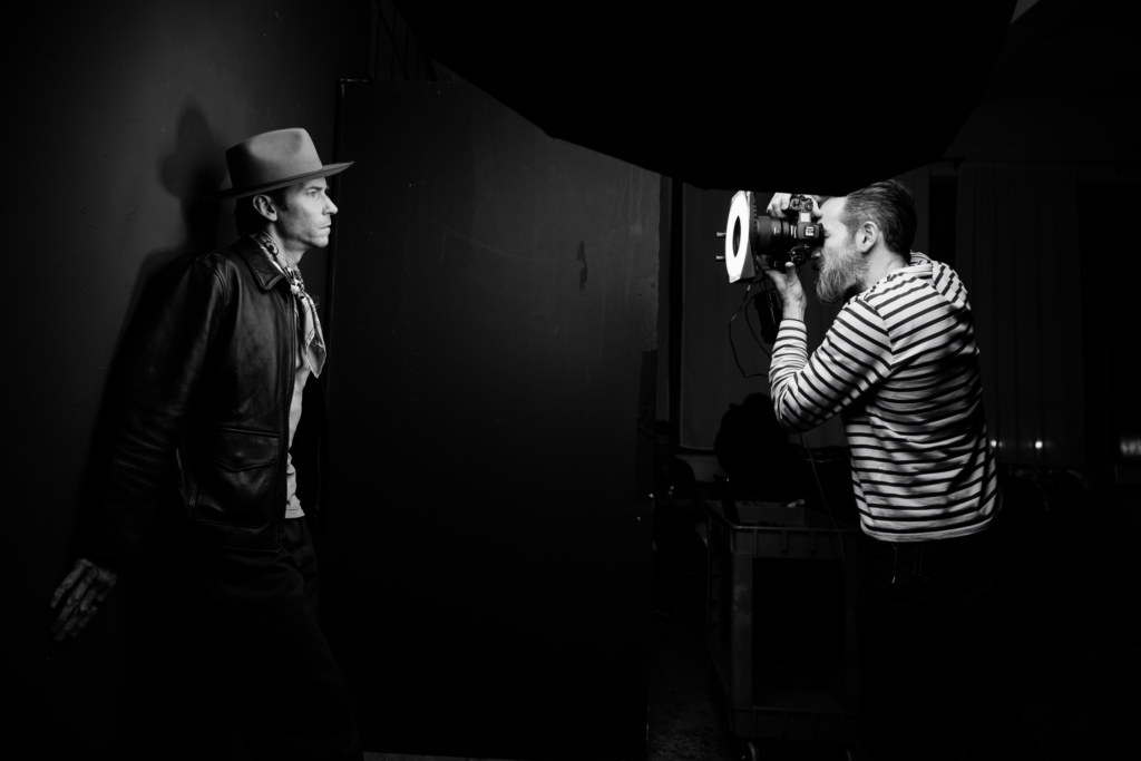 A black-and-white behind-the-scenes photograph of Ivan Weiss working in his studio, leaning forward as he photographs a man standing against a dark wall. A large softbox light hangs above them, shaping the light across her face while he looks through the camera with focused concentration. The scene captures the quiet intensity of a portrait session in progress.

Shot by Rich Soublet.
