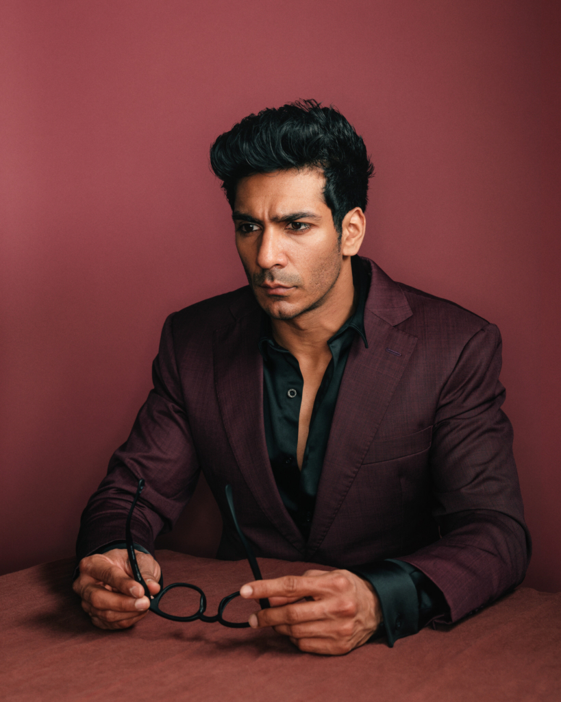 A studio portrait of a man seated at a table against a deep red background. He wears a dark burgundy suit jacket over a black shirt, slightly open at the collar, and holds a pair of eyeglasses loosely in both hands. His gaze is directed off to the side, his expression serious and focused, while the warm, controlled lighting enhances the rich tones of his clothing and backdrop.