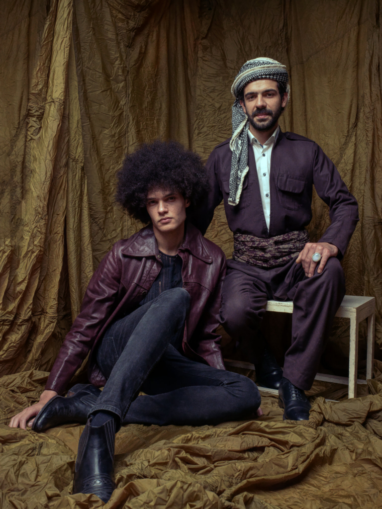 A studio portrait of two men posed against a backdrop of textured golden fabric that drapes onto the floor. One sits on a simple white bench wearing traditional layered garments and a patterned headscarf, while the other sits on the floor beside him in a burgundy leather jacket and dark jeans, his legs extended forward. Both look directly at the camera with calm, steady expressions, the warm, controlled lighting enhancing the rich tones and creating a composed, painterly atmosphere.