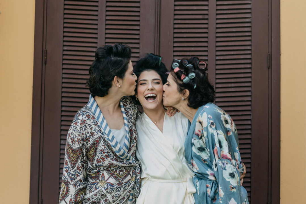 Two women kiss a smiling bride on the cheeks as they stand together in front of a wooden door, sharing a joyful moment.