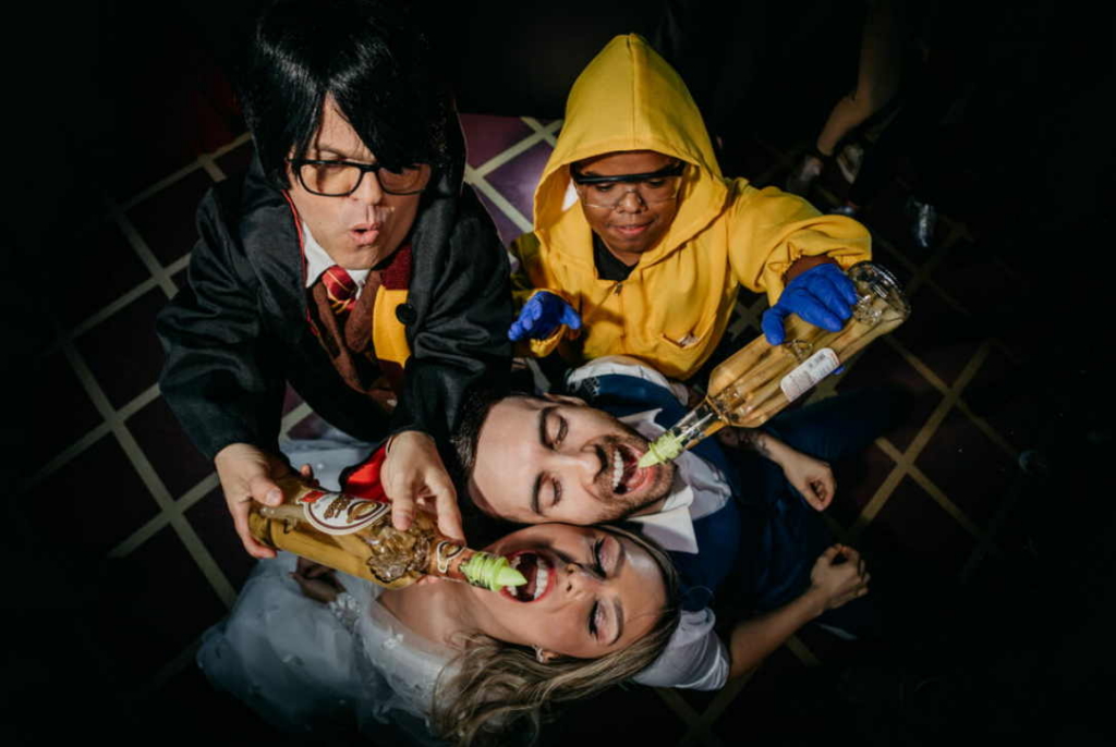 bride and groom lie on the floor while others pour drinks into their mouths, creating a playful and chaotic party scene.
