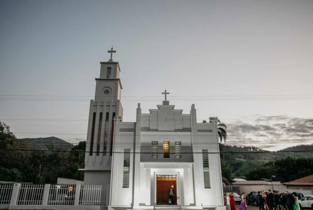 The exterior of a white church is shown at dusk, with people gathered near the entrance.