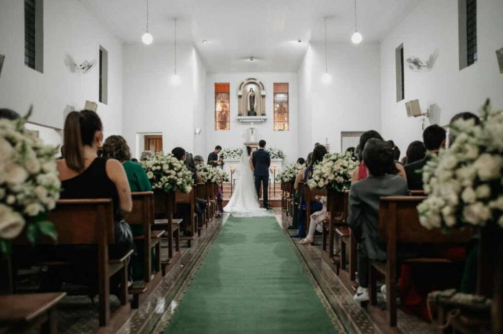 A wedding ceremony takes place inside a church, with guests seated on both sides of the aisle facing the couple at the altar.