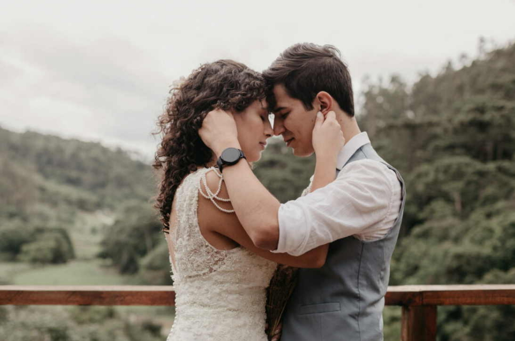 Um casal está em pé, bem próximo um do outro, abraçando-se suavemente com as testas encostadas, cercado por uma paisagem verde e arborizada.