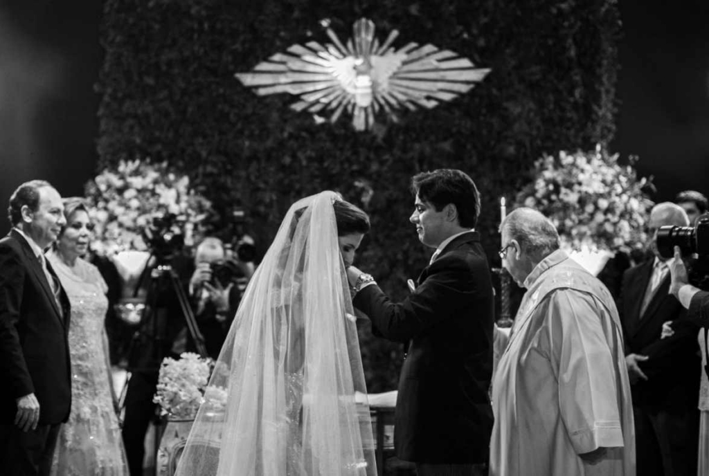 A couple stands at the altar during a formal wedding ceremony as a priest and guests look on in a decorated church.