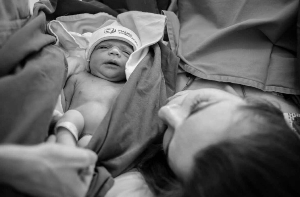 A newborn baby lies wrapped in blankets while an adult looks at them closely in an intimate hospital scene.