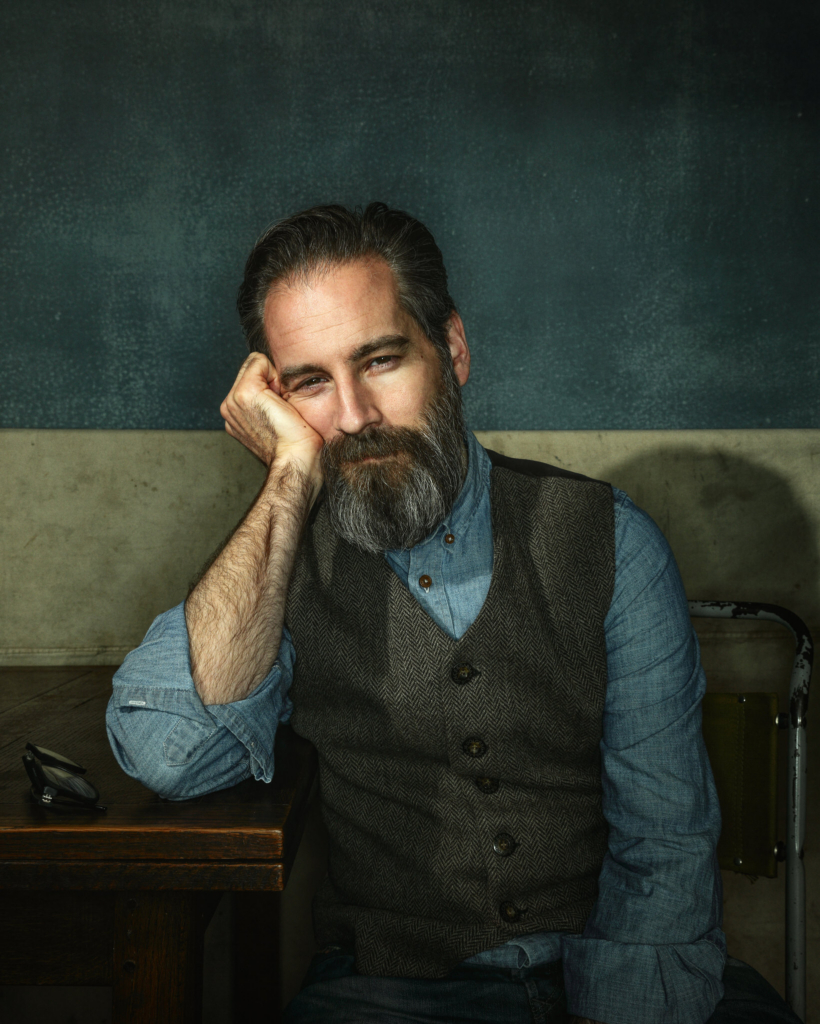A portrait of photographer Ivan Weiss seated at a wooden table, resting his head gently on his hand. He has dark hair streaked with gray and a full salt-and-pepper beard, and he looks calmly and directly at the camera with a thoughtful expression. He wears a blue buttoned shirt and a textured brown vest, set against a softly lit, painterly green backdrop that gives the image a classic, studio portrait feel.