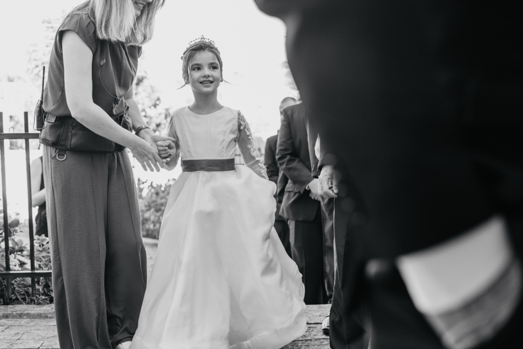 A young girl in a formal dress walks forward holding an adult’s hand during an outdoor ceremony.