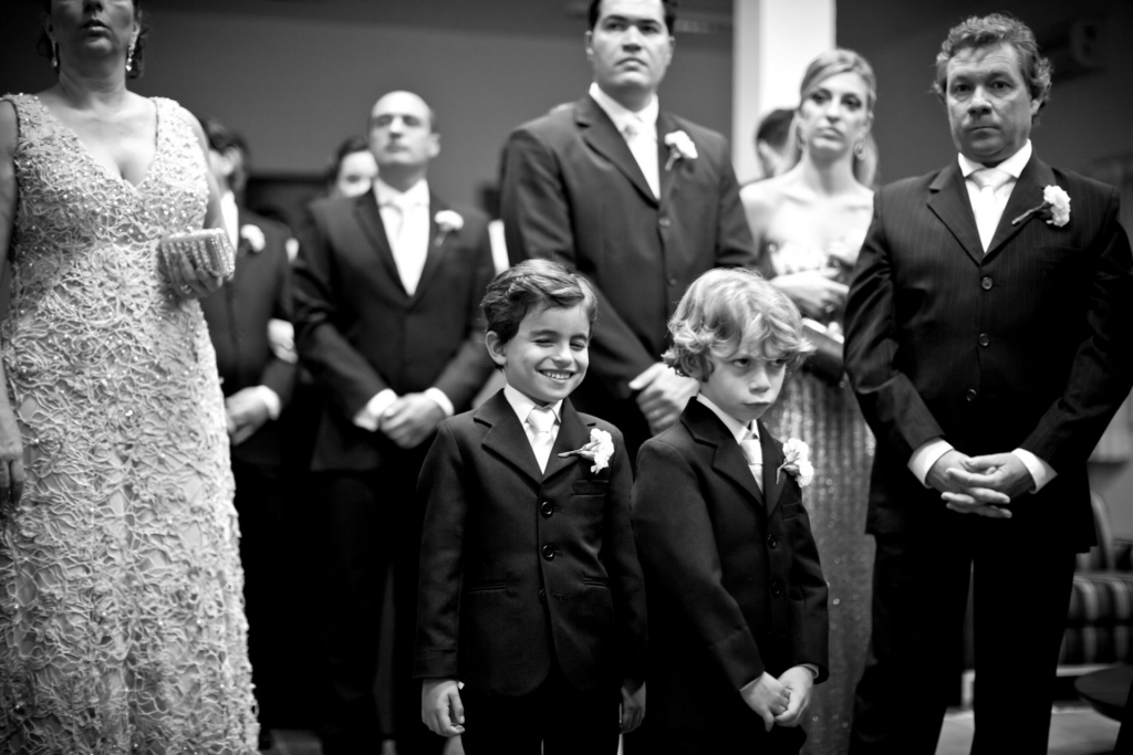 Two young boys in suits stand in front of a group of formally dressed adults, one smiling while the other looks serious.