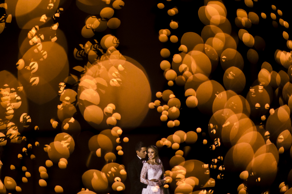 A couple embraces in a dark setting, surrounded by warm, glowing out-of-focus lights.