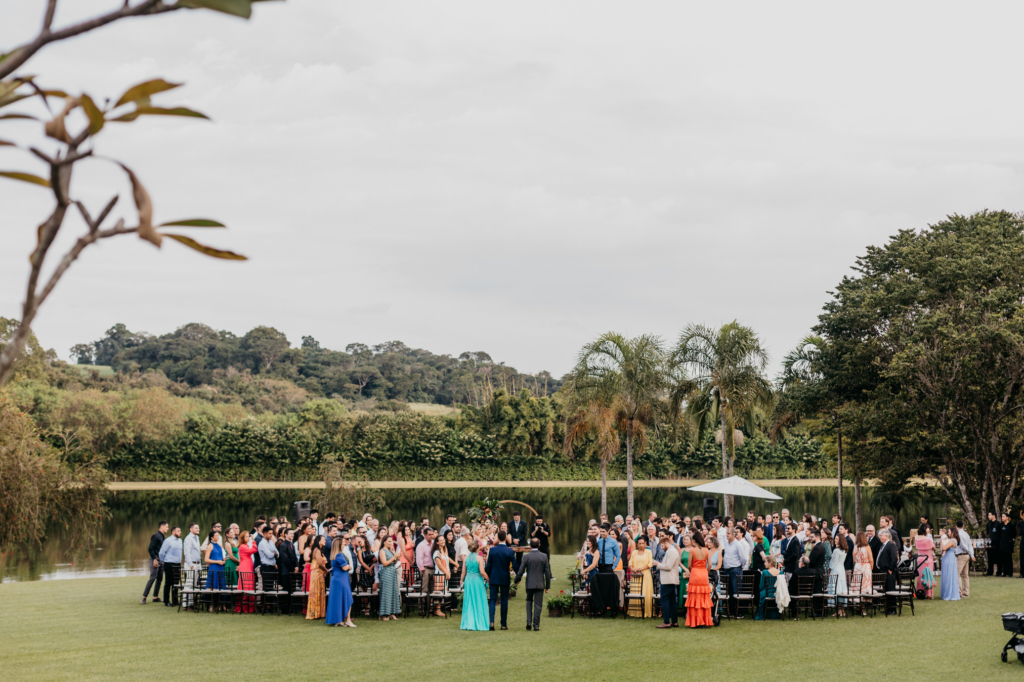 Um grande grupo de pessoas se reúne ao ar livre, sobre um gramado perto de um lago, organizado em círculo para o que parece ser uma cerimônia de casamento, cercado por árvores e vegetação.