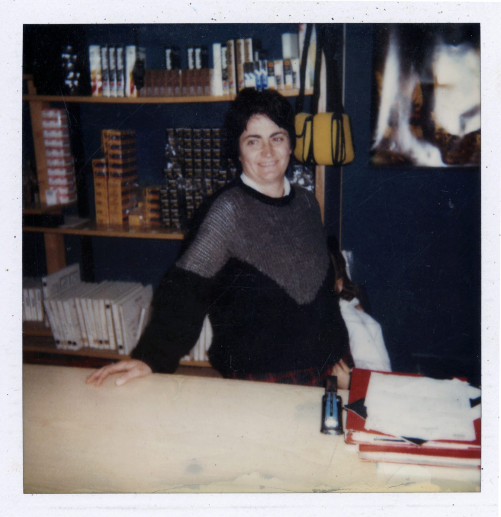 Photographer’s mother standing in the family shop, surrounded by shelves of photo supplies.