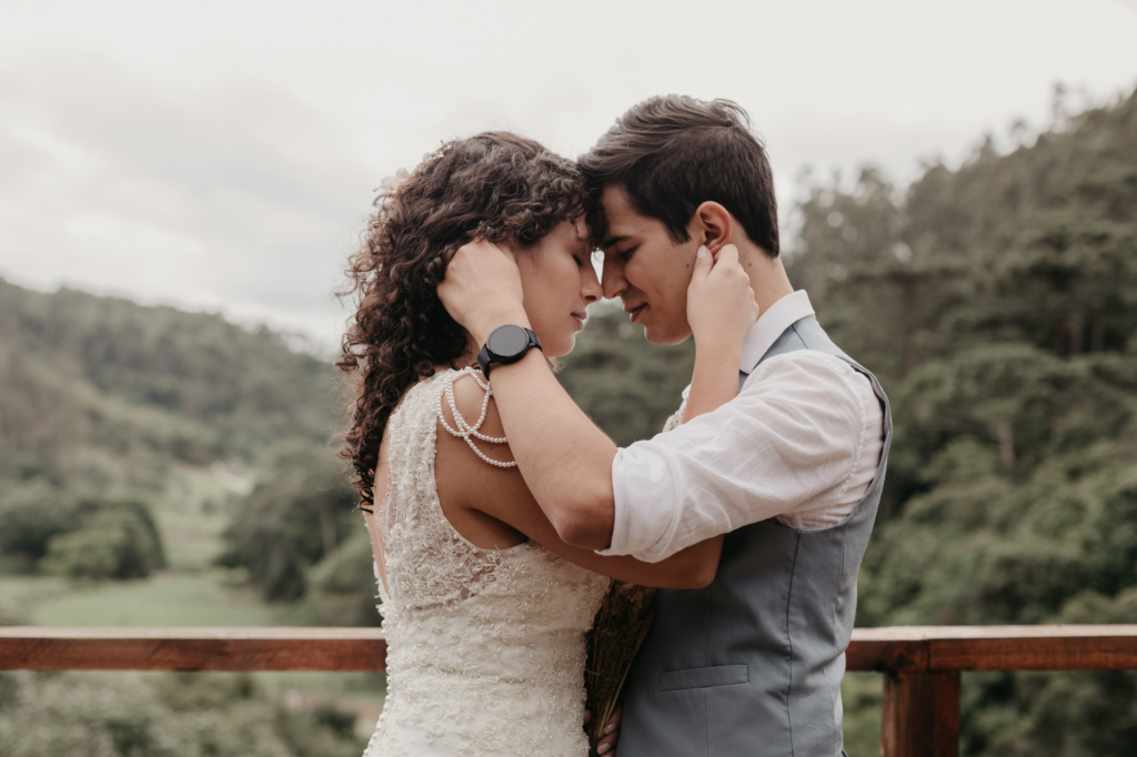 A couple stands close together outdoors, gently holding each other with their foreheads touching, surrounded by a green, forested landscape.