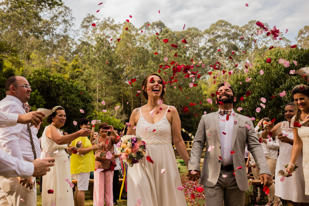 A smiling couple walks hand in hand outdoors while guests throw flower petals around them in celebration.