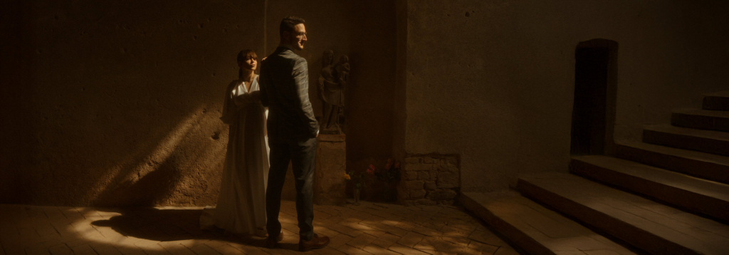 A couple stands together inside a historic stone building, illuminated by a narrow beam of warm light entering from above. The soft light highlights their faces and casts long shadows across the textured walls and floor.

The scene feels quiet and cinematic, with stone columns and a staircase fading into darkness, creating a dramatic and intimate atmosphere.