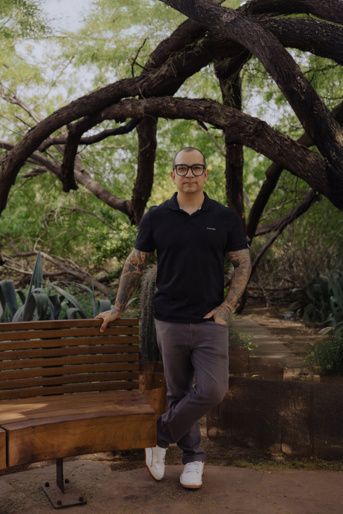 This is a color portrait of photographer Betto Robles standing outdoors in a lush garden. He leans casually against a wooden bench, wearing a black polo shirt and glasses, with tattooed arms visible. Twisted tree branches form an arch behind him, framing his figure against soft green foliage and natural light.