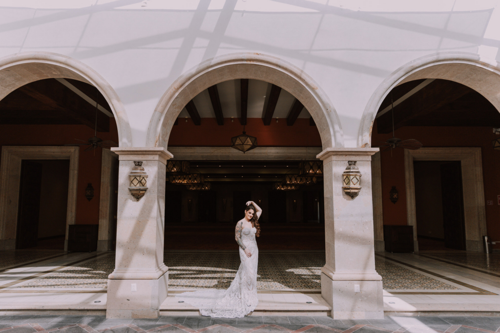 A bride in a fitted white lace gown stands alone beneath a large stone archway, centered within a grand open courtyard.
The architecture dominates the frame with tall white arches, patterned tile floors, and deep shadows, making her appear small and elegant within the space.