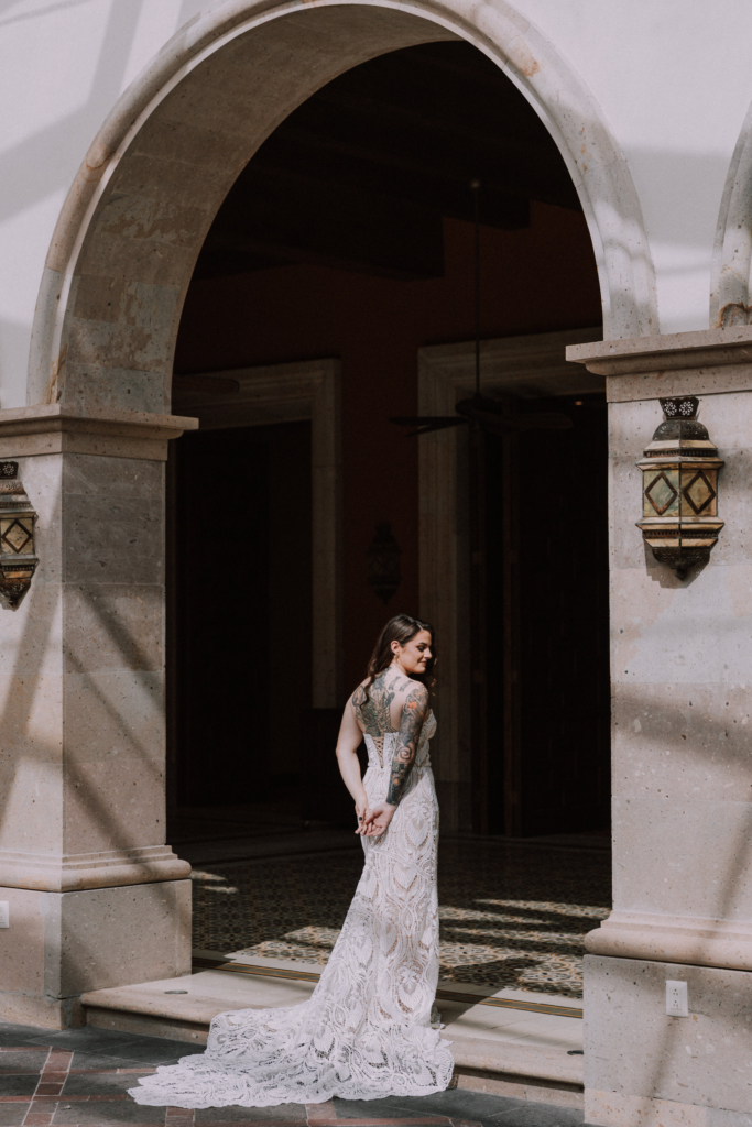 A bride in a fitted lace gown stands beneath a large stone archway, looking over her shoulder.
Soft sunlight highlights her tattoos and the texture of the dress, while the dark interior behind her adds contrast and depth.