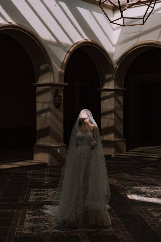 A bride stands alone in a shadowy courtyard beneath tall stone arches, wrapped in a long sheer veil.
Stripes of sunlight fall across the walls and floor, while one bright beam lights her face and gives the portrait a quiet, dramatic mood.