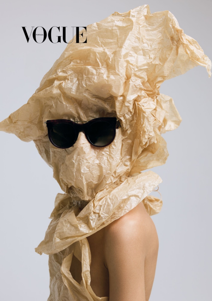 A fashion image of a figure wrapped in crumpled beige paper, wearing black sunglasses against a clean studio background. The look feels playful and sculptural, turning simple materials into something theatrical.