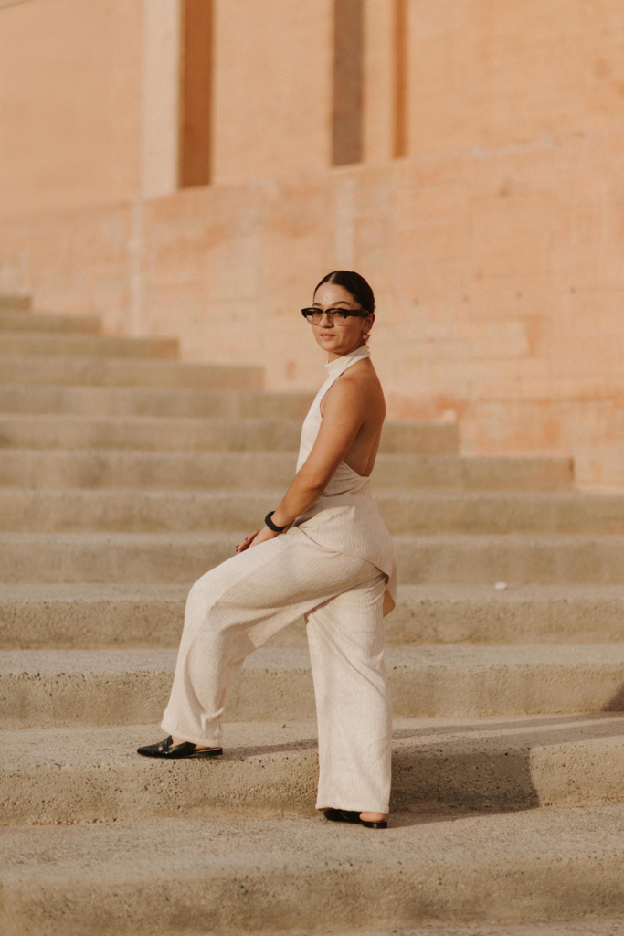 This is a color portrait of photographer Mayra Mungarro standing on wide stone steps in warm, golden light. She wears a light, sleeveless outfit and sunglasses, turning her body slightly while looking toward the camera. The soft beige tones of the architecture and the sunlight create a calm, minimal backdrop around her.