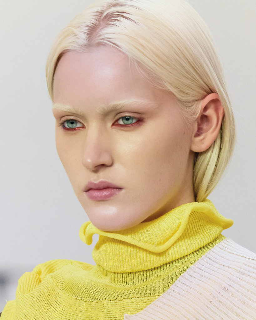 A close-up portrait of a model with pale blonde hair and minimal eyebrows, wearing a soft yellow high-neck garment. The image is clean and precise, with a focus on skin, color, and subtle expression.