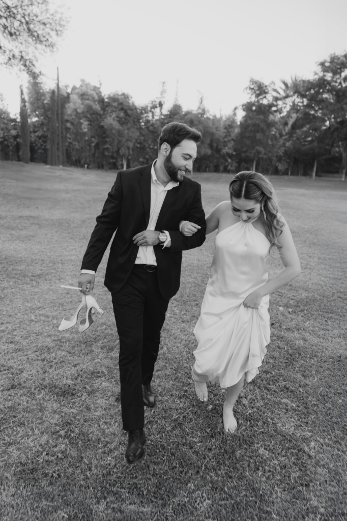 This is a black-and-white photograph of a couple walking together across an open grassy field. The man holds a pair of high heels in one hand while the woman walks barefoot, lifting her dress slightly as they move. Both are smiling and looking down, sharing a relaxed, playful moment.