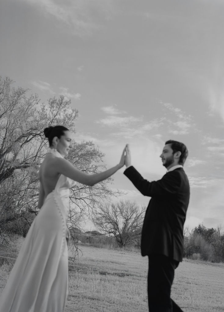 This is a black-and-white photograph of a couple standing outdoors, facing each other with their hands pressed together. The bride wears a long dress, and the groom is dressed in a suit, both slightly blurred as if in motion. Bare trees and an open landscape stretch behind them under a wide, quiet sky.