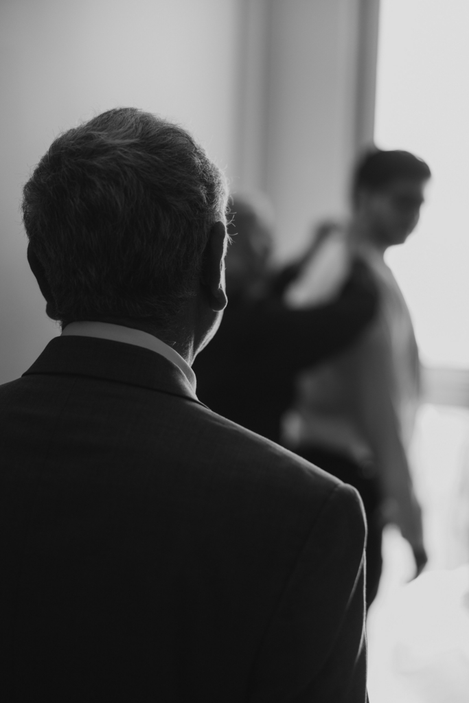 This is a black-and-white photograph taken from behind an older man, his figure in sharp focus as he looks ahead. In the distance, a younger man stands near a window while another figure adjusts his clothing, both softly blurred. The contrast in focus creates a sense of observation and quiet anticipation.