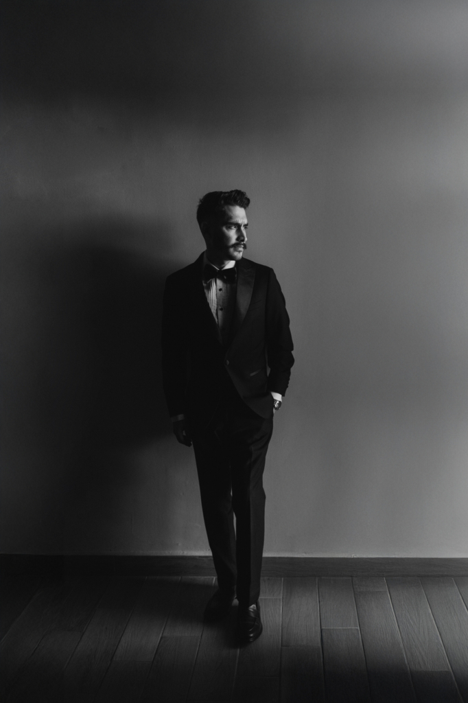 This is a black-and-white portrait of a man in a suit standing against a plain wall. He looks off to the side, one hand in his pocket, as light falls across his face and body, casting a long shadow beside him. The minimal setting and strong contrast create a quiet, cinematic mood.