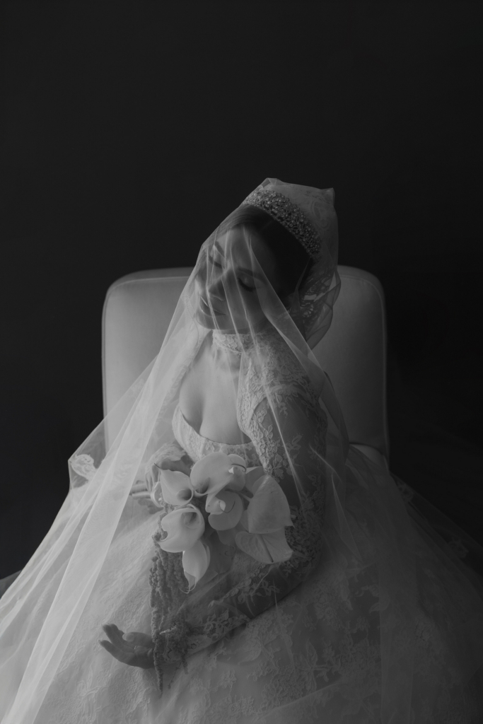 This is a black-and-white portrait of a bride seated in a chair, softly lit against a dark background. She wears a lace gown and a veil that drapes over her face, holding a bouquet close to her chest. Her eyes are closed and her head tilts gently, creating a quiet, intimate and almost dreamlike mood.