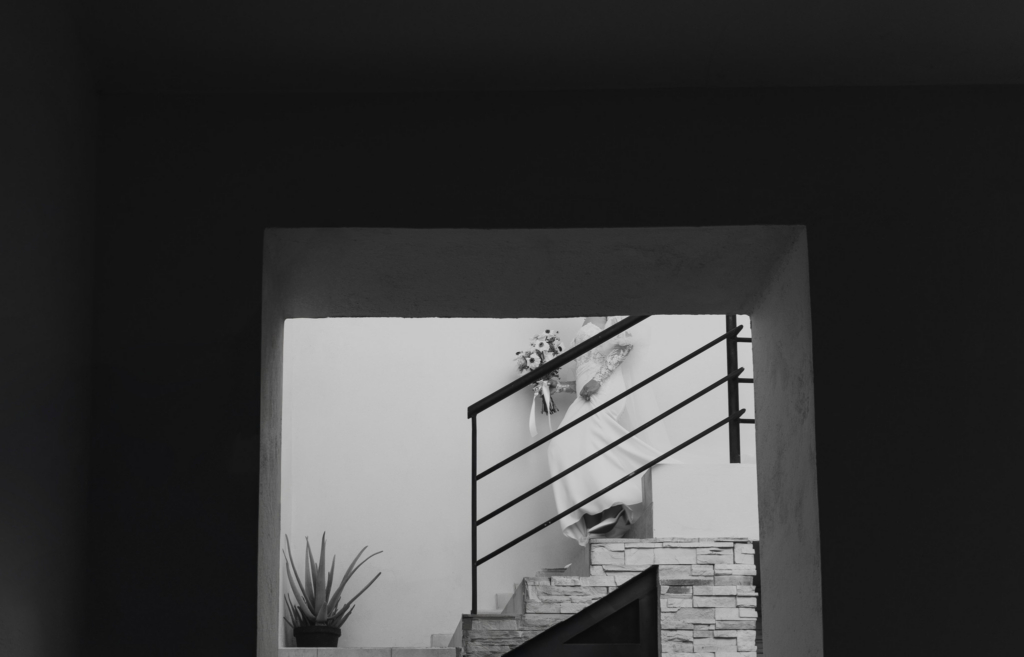 This is a black-and-white photograph framed through a dark architectural opening, creating a strong vignette around the scene. A bride in a flowing dress is seen mid-step on a staircase, holding a bouquet, partially obscured by the railing. The composition emphasizes contrast between the bright interior and the deep shadows surrounding it.