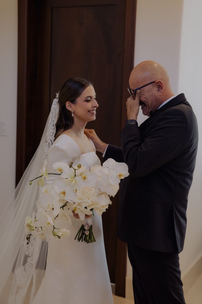 This is a color photograph of a bride standing indoors with an older man, likely her father. She holds a bouquet of white flowers and smiles gently, while he wipes tears from his eyes, visibly emotional. The soft light and simple background focus attention on their intimate, tender moment.