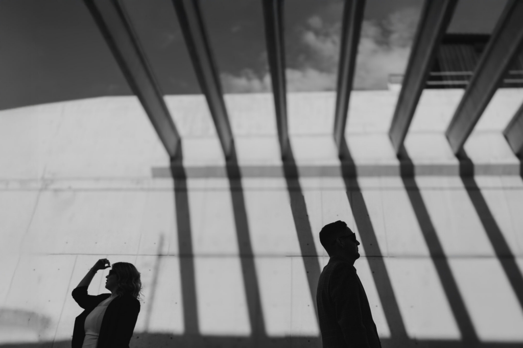 This is a black-and-white photograph of two people standing apart, facing opposite directions in a large architectural space. Strong vertical shadows from overhead beams stretch across the wall, creating bold graphic lines around them. Their figures appear as dark silhouettes, emphasizing distance and tension within the composition.