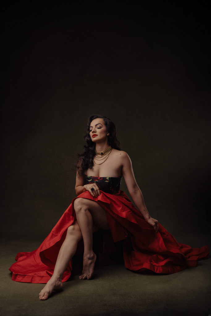 This is a color portrait of a woman seated on a low chair, wearing a dramatic red skirt and a strapless top with floral details. She sits barefoot with one leg crossed, her eyes closed and head slightly tilted, creating a calm, composed presence. The dark background and soft lighting emphasize the richness of the red fabric and her sculptural pose.