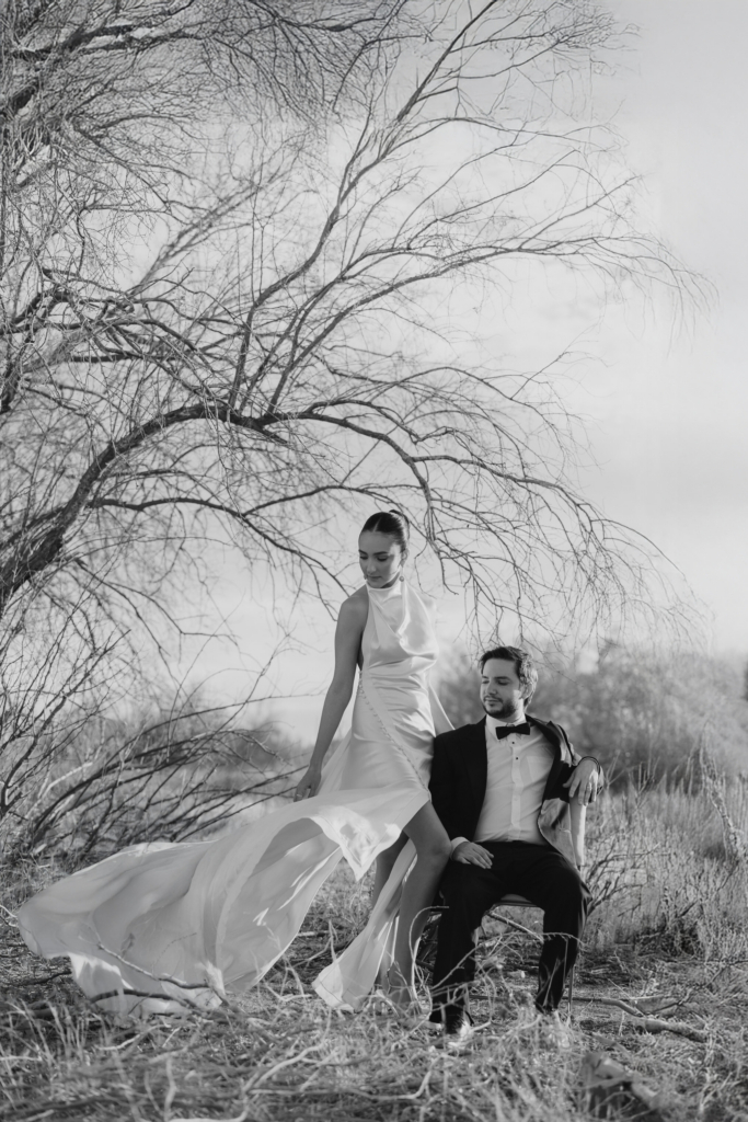 This is a black-and-white photograph of a couple posed outdoors beneath a large, bare tree. The woman stands beside a seated man, her dress flowing in the wind as she looks downward, while he looks up at her. The branches spread widely above them, adding a dramatic, sculptural backdrop to the scene.
