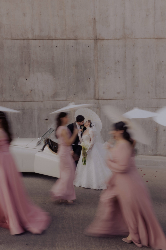 This is a color photograph of a bride and groom standing close together beside a white vintage car. They share a quiet moment, leaning in toward each other, while blurred figures in flowing dresses move past them in the foreground. The motion around them contrasts with their stillness, drawing focus to their intimate connection.