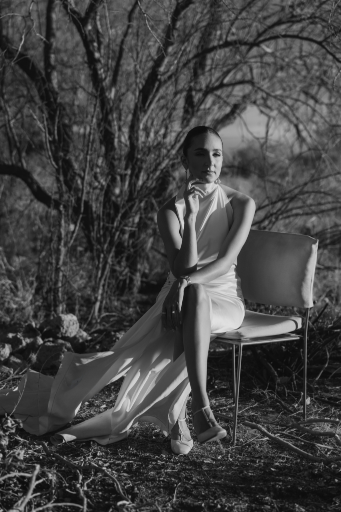 This is a black-and-white portrait of a woman seated on a chair outdoors among bare trees and dry ground. She wears a long, flowing dress and sits with one leg crossed, resting her hand thoughtfully near her face. The light creates strong contrast across her figure, giving the scene a quiet, sculptural feel.