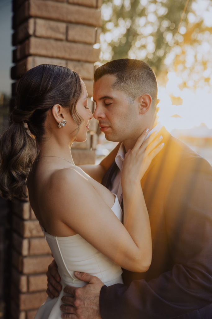 This is a warm, close-up color photograph of a couple embracing outdoors at sunset. Their faces are close, eyes gently closed, as soft golden light and lens flare wrap around them. The scene feels intimate and quiet, with the brick wall and blurred background framing the moment.