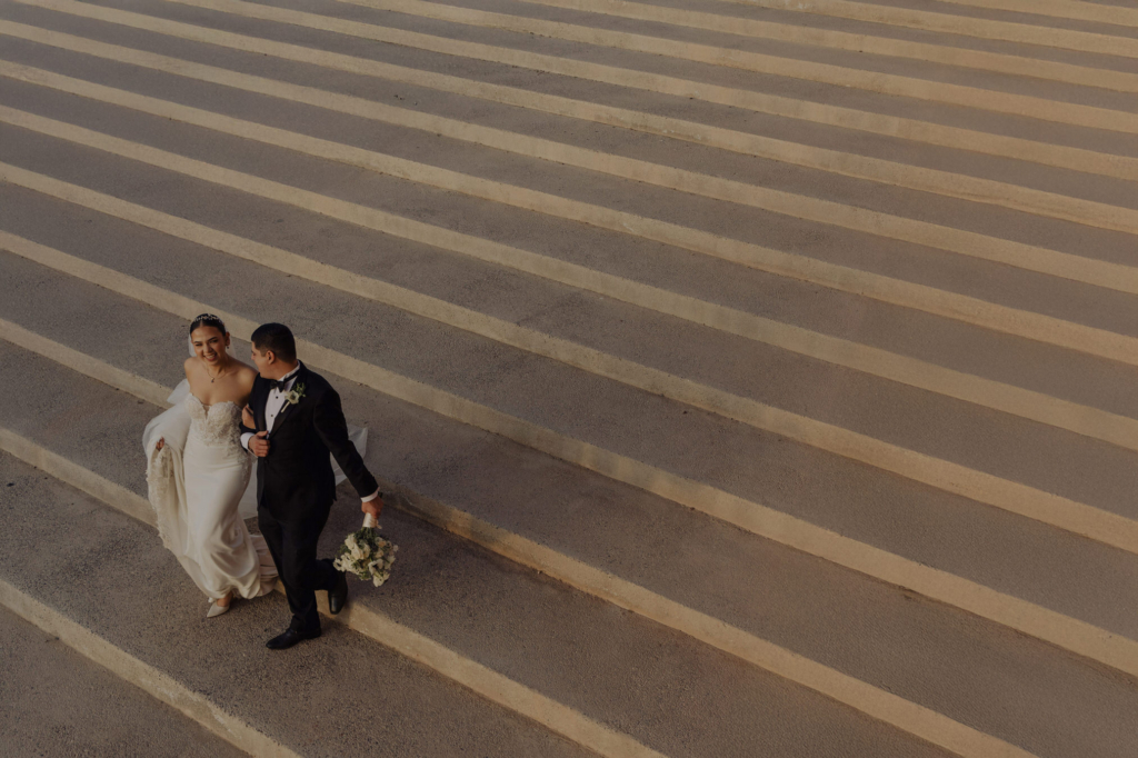 This is a color photograph taken from above, showing a bride and groom walking together across wide, patterned steps. They move side by side, smiling at each other, with the groom holding a bouquet. The repeating lines of the steps create a strong graphic rhythm that contrasts with their intimate moment.
