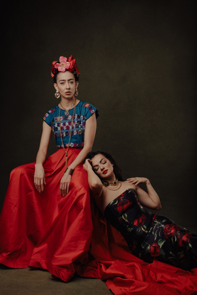 This is a color portrait of two women posed against a dark, textured background. One sits upright in a vivid red skirt and embroidered top with a floral headpiece, while the other reclines beside her in a black dress with red patterns. Their contrasting poses and rich colors create a dramatic, painterly composition.