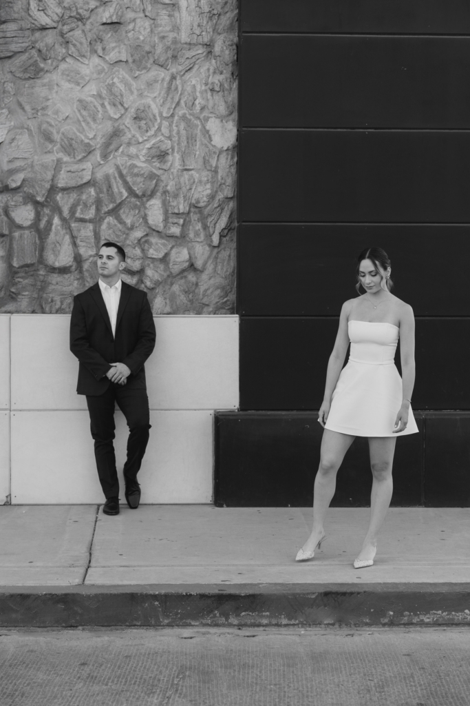 This is a black-and-white photograph of a couple standing apart against a contrasting wall of stone and dark panels. The man leans back with his hands together, looking off to the side, while the woman stands a few steps away, looking down. The composition emphasizes distance between them through space, posture, and the stark geometric background.