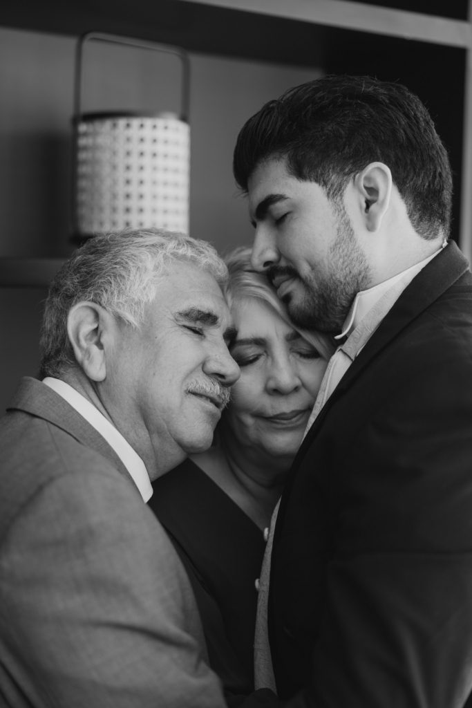 This is a black-and-white close-up of three people embracing closely, likely family members. Their faces rest gently against one another, eyes closed, sharing a quiet, emotional moment. The tight framing and soft light emphasize intimacy and connection.