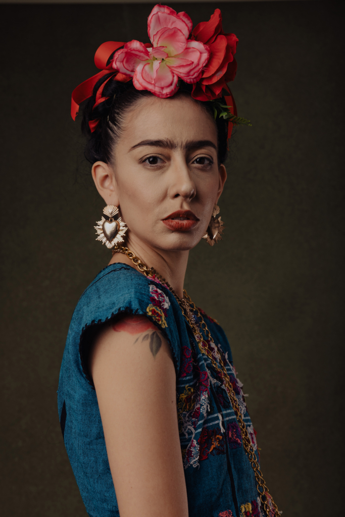 This is a color portrait of a woman inspired by Frida Kahlo, wearing a floral headpiece with red ribbons and bold jewelry. She faces the camera with a direct, steady gaze, her expression calm and self-possessed. The rich colors of her embroidered dress contrast with the dark, textured background, drawing attention to her face and adornments.