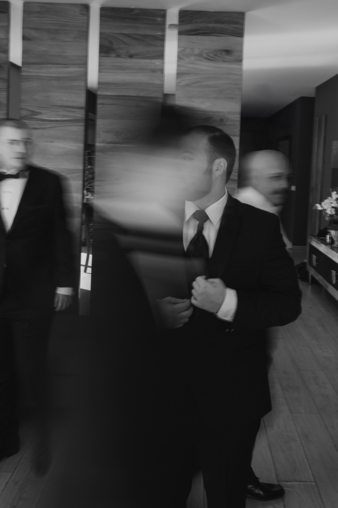 This is a black-and-white photograph of men in suits moving through an interior space. One man stands in partial focus adjusting his jacket, while others pass by as blurred figures. The motion blur creates a sense of urgency and preparation within the scene.