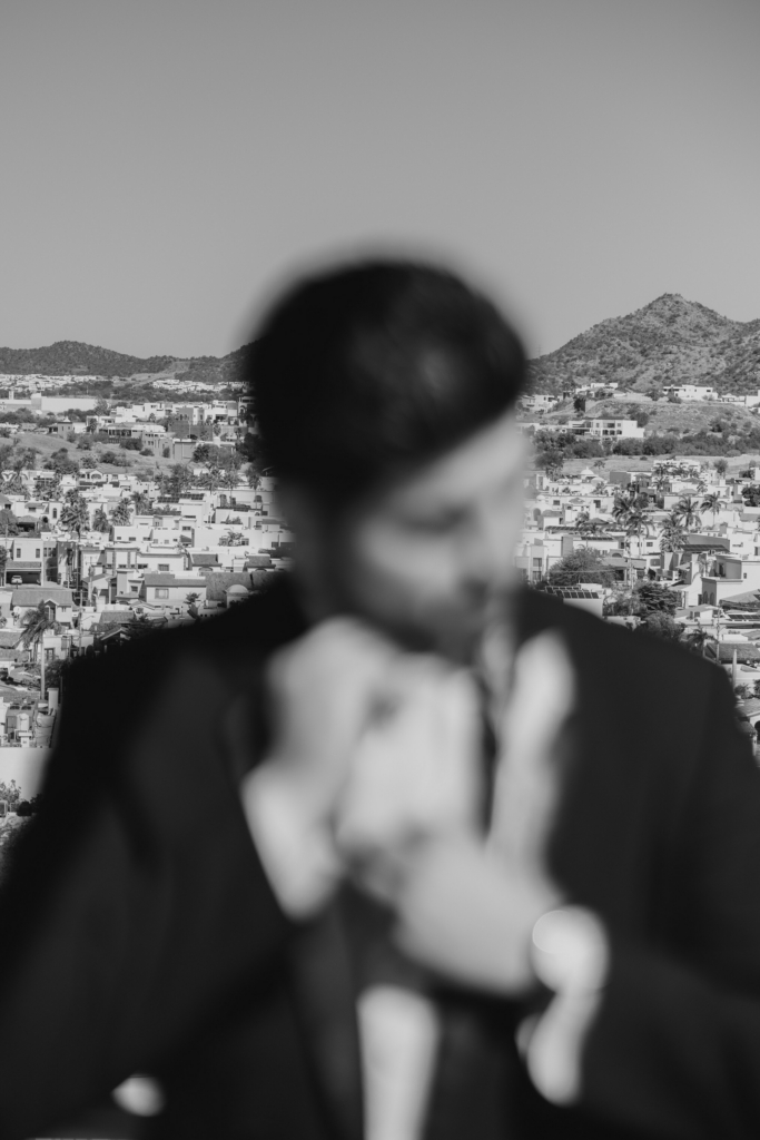 This is a black-and-white photograph with a blurred figure of a man in the foreground, adjusting his suit. Behind him, a sharp cityscape stretches across the frame, with houses and hills in the distance. The contrast between the soft, out-of-focus subject and the detailed background creates a layered, disorienting effect.