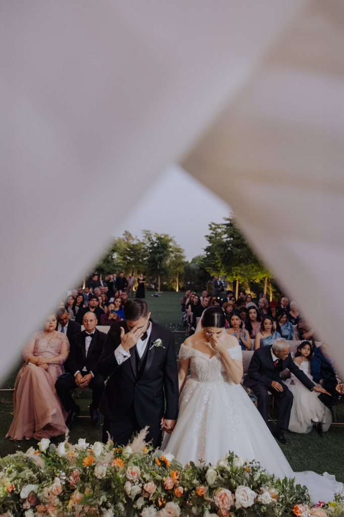 This is a color photograph of a bride and groom standing at the front of an outdoor ceremony, framed by soft fabric in the foreground. Both cover their faces with their hands, appearing emotional as they stand side by side holding hands. Guests sit behind them, watching quietly, while floral arrangements fill the lower part of the scene.