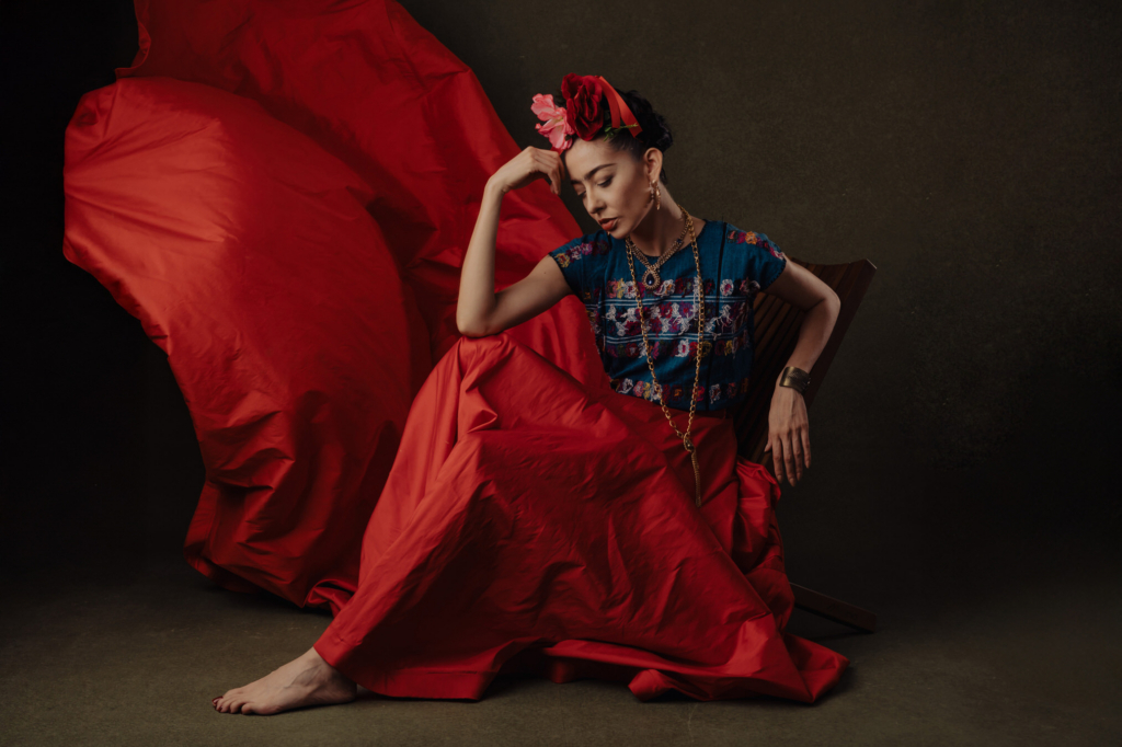 This is a color portrait of a woman seated on the floor, wrapped in a dramatic, flowing red skirt that spreads widely behind her. She wears a blue embroidered top, gold jewelry, and a floral headpiece, with her head tilted down in a quiet, introspective pose. The lighting is soft and controlled, emphasizing the rich textures of the fabric and the contrast between deep red and muted background tones.