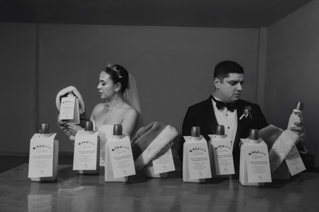 This is a black-and-white photograph of a bride and groom standing behind a table lined with identical bottles and folded blankets. Each of them examines an item separately, turned slightly away from one another. The repeated objects create a rhythmic pattern, while their opposing gazes suggest quiet separation.